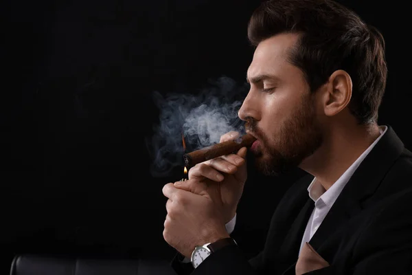 Handsome man lightning cigar on black background. Space for text