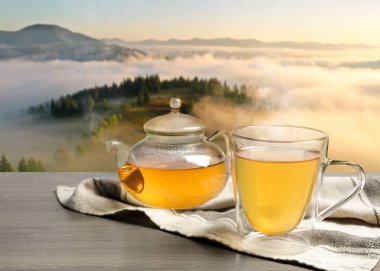 Cam bardakta yeşil çay ve sisli dağlarda ahşap masada çaydanlık.