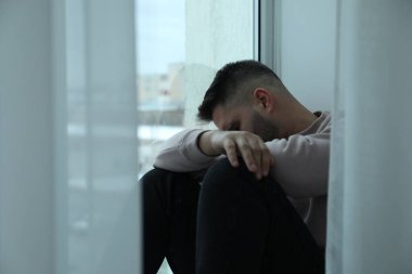 Evdeki pencerenin yanında oturan üzgün adam.