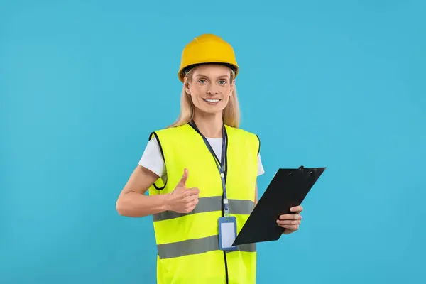 Engineer in hard hat holding clipboard and showing thumb up on light blue background