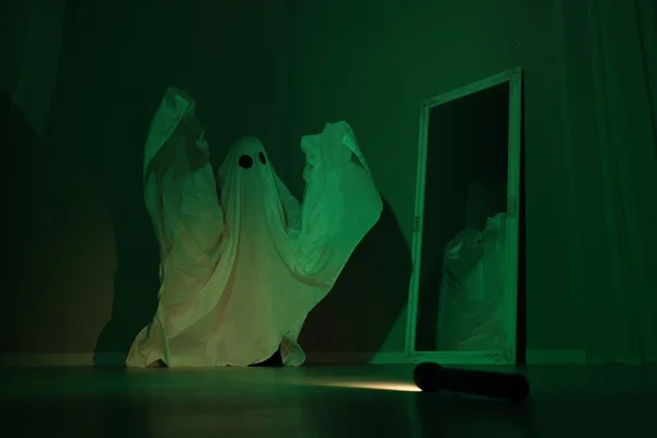Creepy ghost. Woman covered with sheet near mirror in green light
