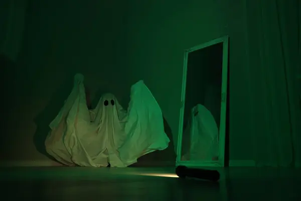 Creepy ghost. Woman covered with sheet near mirror in green light