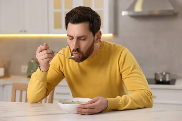 Man eating delicious chicken soup at light marble table in kitchen