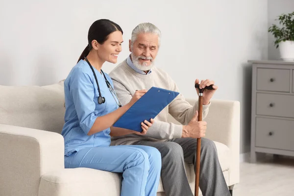 Smiling nurse with clipboard assisting elderly patient on sofa in hospital