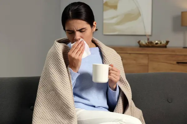 Woman with tissue and cup of drink coughing on sofa at home. Cold symptoms