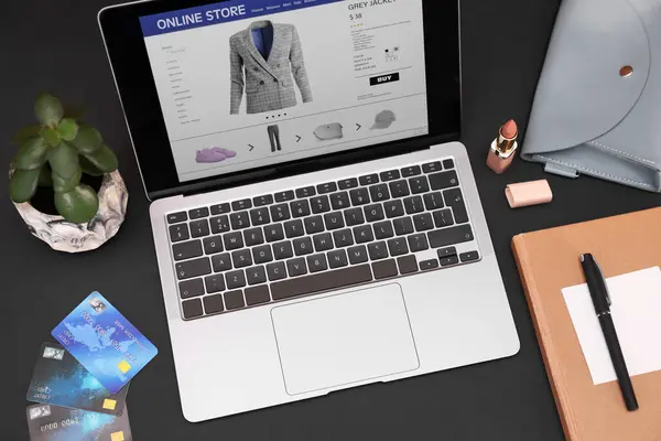Online store website on laptop screen. Computer, credit cards, women\'s bag, stationery and lipstick on black background, above view
