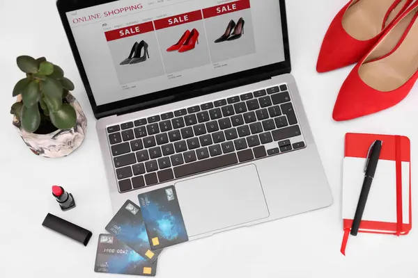 Online store website on laptop screen. Computer, credit cards, women\'s shoes, stationery and lipstick on white background, above view