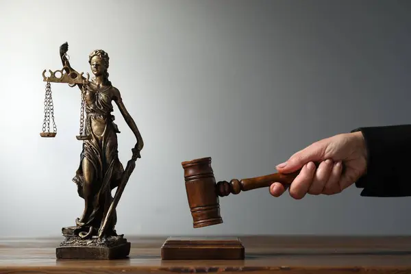Judge striking mallet at wooden table against grey background, closeup. Figure of Lady Justice indoors