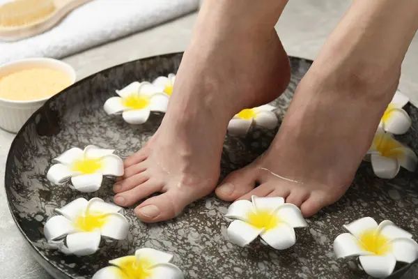 Woman soaking her feet in bowl with water and flowers on floor, closeup. Spa treatment