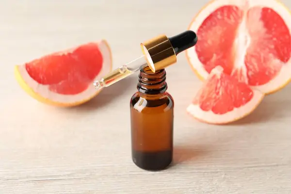Grapefruit essential oil in bottle, pipette and fruit on light wooden table, closeup