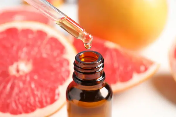 Dripping grapefruit essential oil from pipette into bottle at white table, closeup