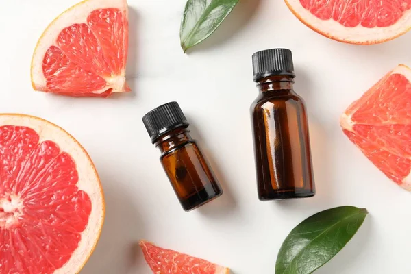 Grapefruit essential oil in bottles, leaves and cut fruit on white table, flat lay