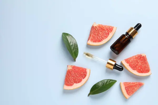 Grapefruit essential oil in bottle, pipette, leaves and cut fruit on light blue table, flat lay. Space for text