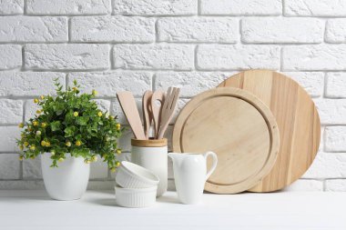 Kesme tahtaları, mutfak aletleri, bulaşık takımları ve ev bitkileri tuğla duvarın yanındaki beyaz masada.