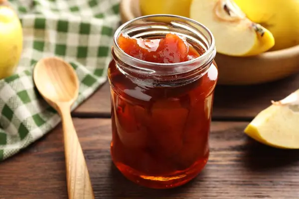 Tasty homemade quince jam in jar, spoon and fruits on wooden table, closeup