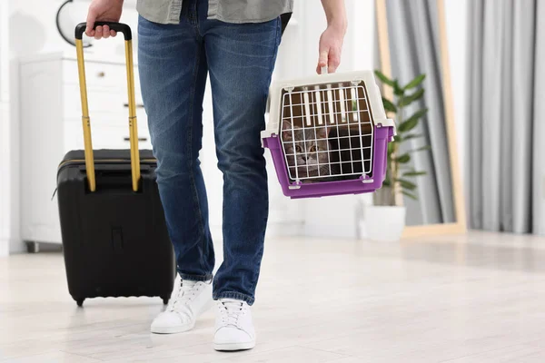 Travel with pet. Man holding carrier with cute cat and suitcase at home