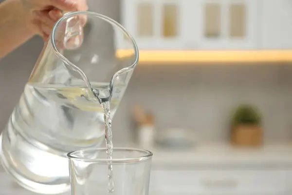 Woman pouring water from jug into glass in kitchen, closeup. Space for text