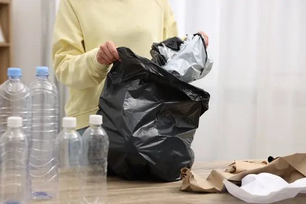 Woman with plastic bag separating garbage at table in room, closeup