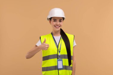 Başlığı ve rozeti olan mühendis bej arka planda baş parmağını gösteriyor.