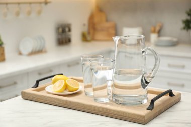 Sürahi, temiz suyla bardaklar ve mutfaktaki beyaz masada limonlar.