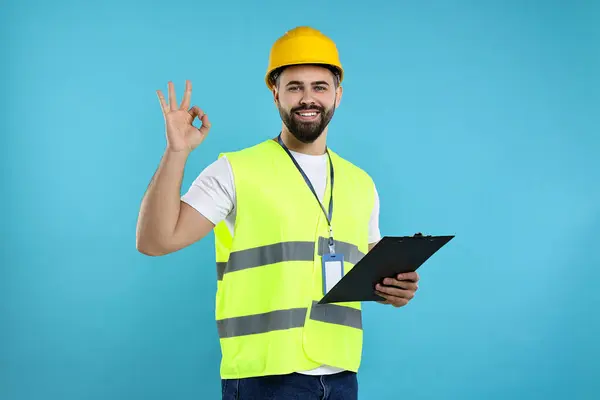Engineer in hard hat holding clipboard and showing ok gesture on light blue background