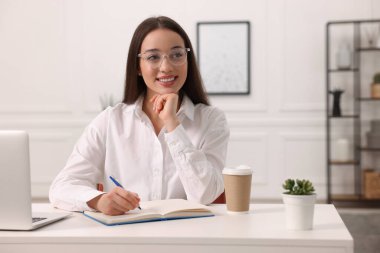 Kapalı alandaki beyaz masada deftere yazan genç bir kadın.
