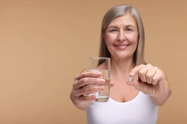 Beautiful woman with vitamin capsule and glass of water on beige background, selective focus. Space for text