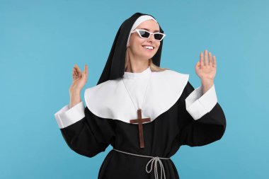 Mavi arka planda güneş gözlüklü, rahibe kıyafetli mutlu kadın.