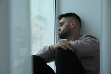 Evdeki pencerenin yanında oturan üzgün adam.