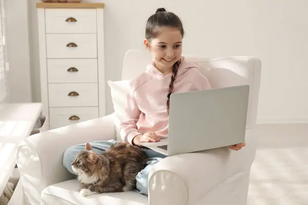 Cute little girl with laptop and cat in armchair at home. First pet