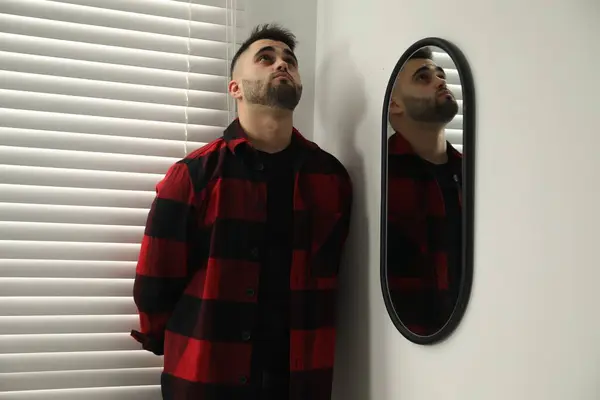 Sad young man near mirror at home