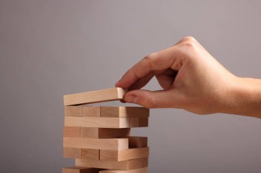 Jenga oynuyorum. Gri arka planda tahta blokları olan kule inşa eden adam, yakın plan.