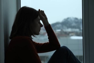 Evdeki pencerenin kenarında oturan üzgün genç kadın.
