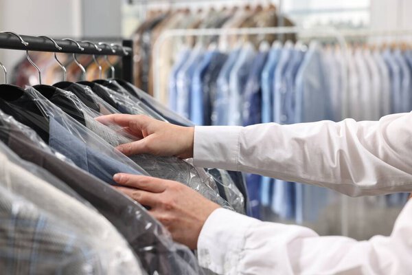 Dry-cleaning service. Woman taking jacket in plastic bag from rack indoors, closeup
