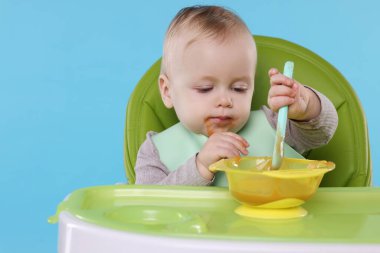 Küçük şirin bebek, açık mavi arka planda, yüksek sandalyede sağlıklı yiyecekler yiyor.