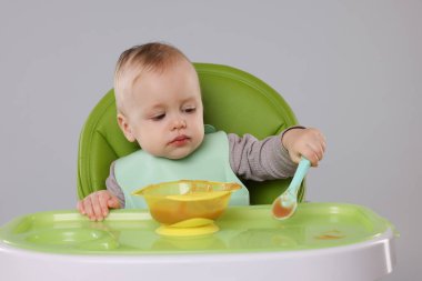Gri arka planda, yüksek sandalyede sağlıklı yiyecekler yiyen şirin bir bebek.