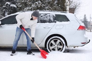 Adam dışarıda arabanın yanında kürekle kar kaldırıyor.