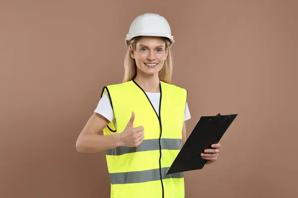 Engineer in hard hat holding clipboard and showing thumb up on brown background