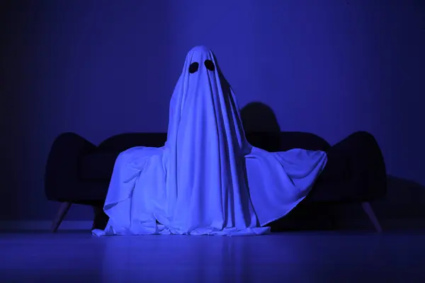 Creepy ghost. Woman covered with sheet sitting on sofa in blue light