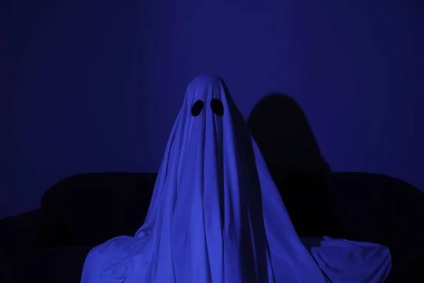 Creepy ghost. Woman covered with sheet sitting on sofa in blue light