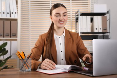 Ofisteki tahta masada dizüstü bilgisayar kullanırken not alan mutlu kadın.