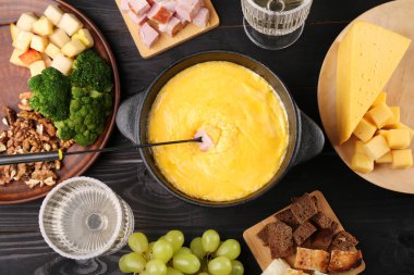 Eritilmiş peynirli fondü tenceresi, çatal ve siyah ahşap masa üzerinde farklı ürünler.