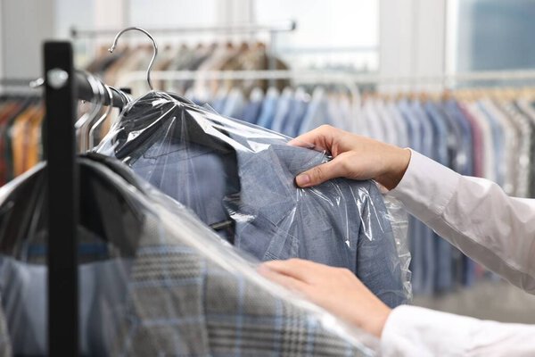 Dry-cleaning service. Woman taking jacket in plastic bag from rack indoors, closeup
