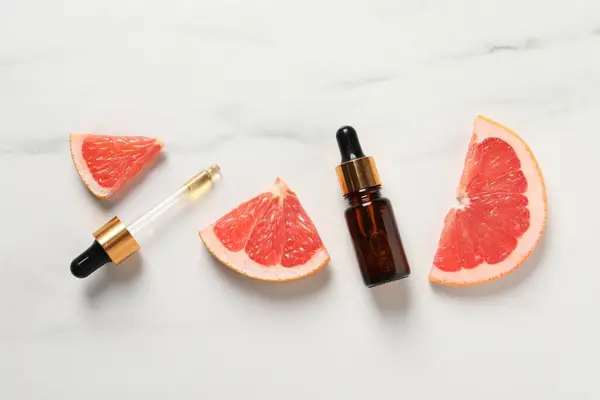 Grapefruit essential oil in bottle, pipette and cut fruit on white marble table, flat lay