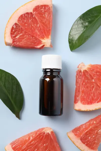 Grapefruit essential oil in bottle, leaves and cut fruit on light blue table, flat lay