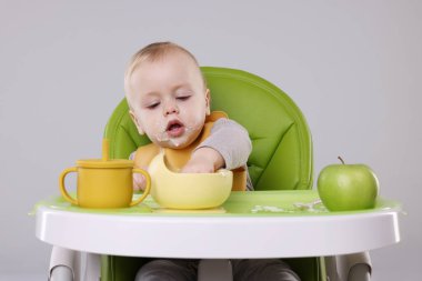 Gri arka planda, yüksek sandalyede sağlıklı yiyecekler yiyen şirin bir bebek.