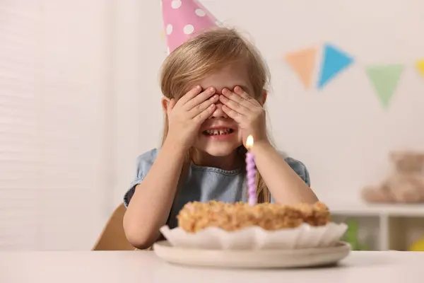 Cute girl in party hat with birthday cake at table indoors
