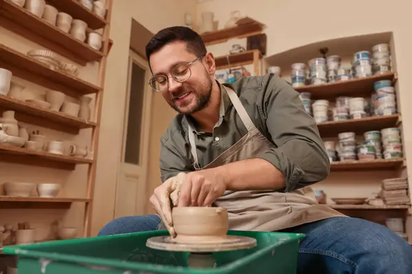 Clay crafting. Happy man making bowl on potter\'s wheel in workshop, low angle view