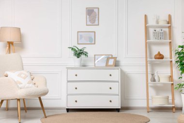 Beyaz duvarda asılı resim çerçeveleri, oturma odasında çekmeceler ve koltuk.