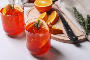 Aperol spritz kokteyli, biberiye ve portakal dilimleri beyaz ahşap masada.